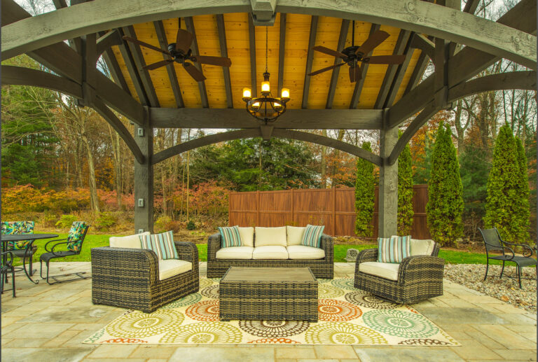 Interior of timber pavilion with arched knee braces and natural tongue-and-groove ceiling, featuring outdoor fans, chandelier, and wicker seating on a stone patio in Westport, Connecticut.