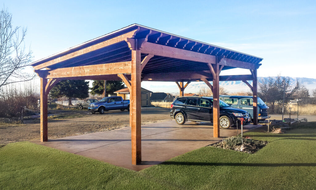 Timber frame cabana-style carport with pitched roof, stained rough-sawn timbers, and open sides sheltering two parked vehicles in a rural setting.