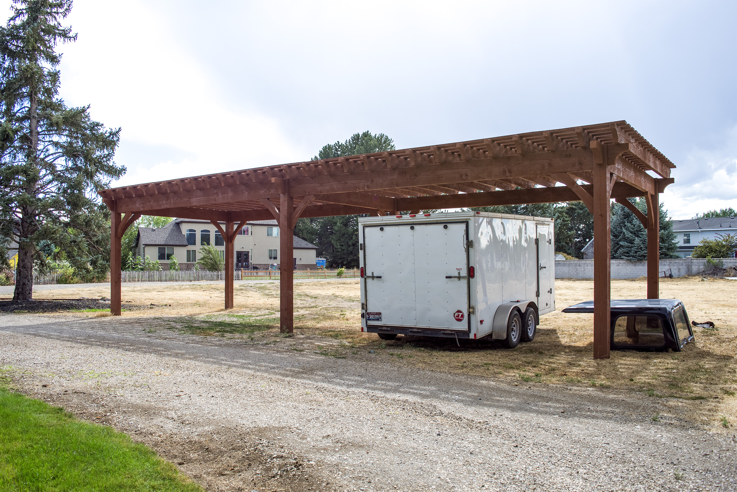 Timber frame carport–pergola with open lattice roof, stained rough-sawn timbers, and curved braces sheltering a white enclosed farm trailer.