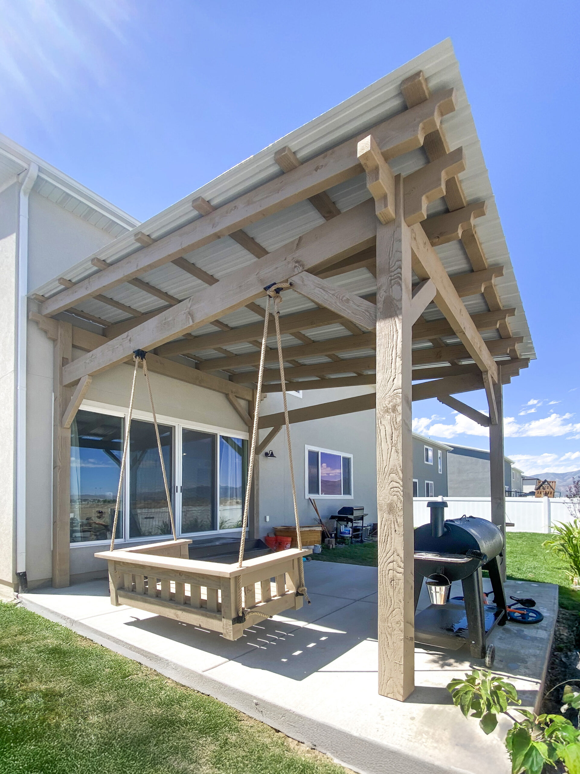 Side view under a timber cabana showing rafters, purlins, and corrugated metal roof above a patio swing.