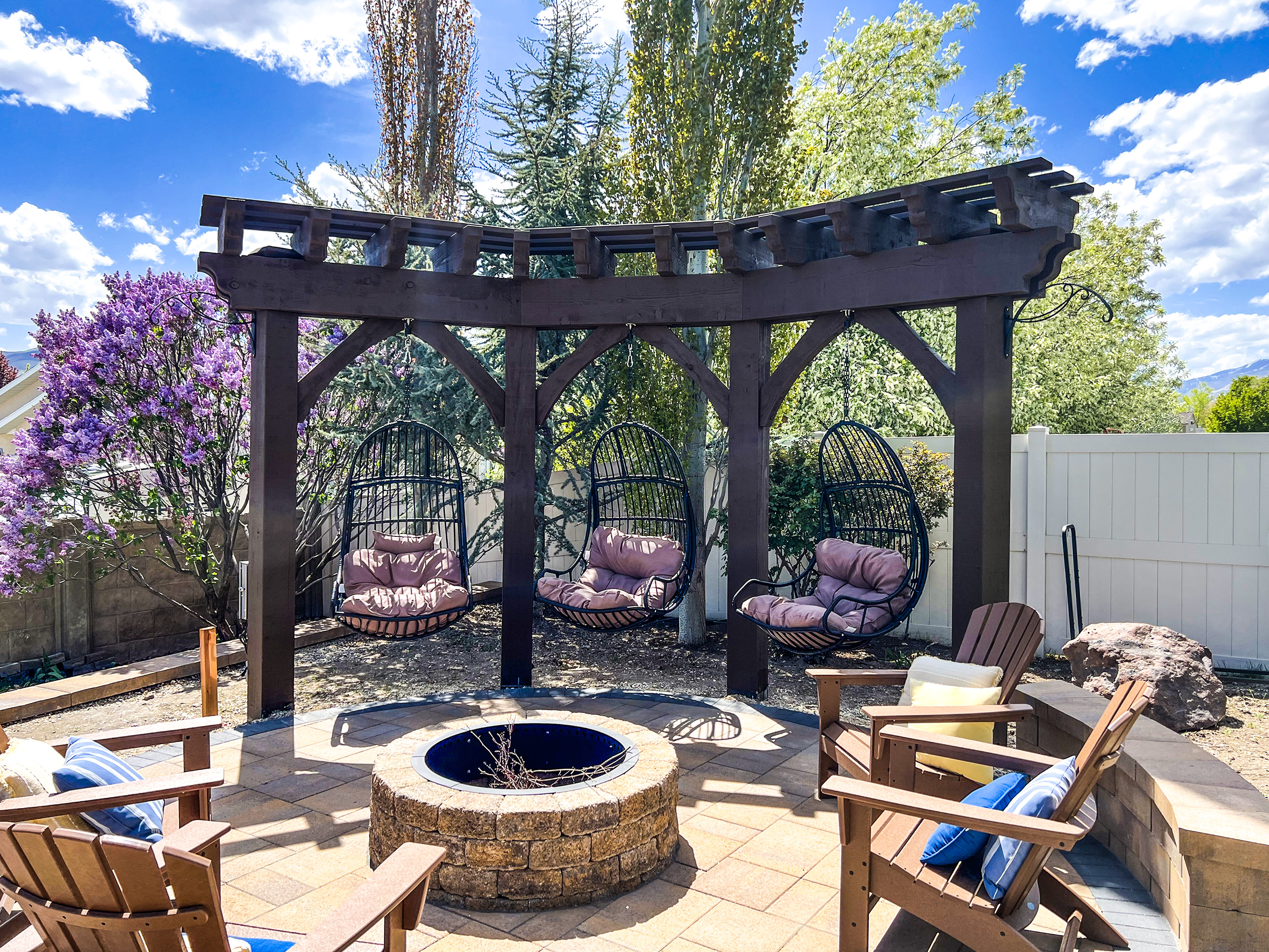 Centered view of timber trellis with curved top beam, hanging egg swings, Rich Cordoba stain, and fire pit patio with Adirondack chairs and garden landscaping
