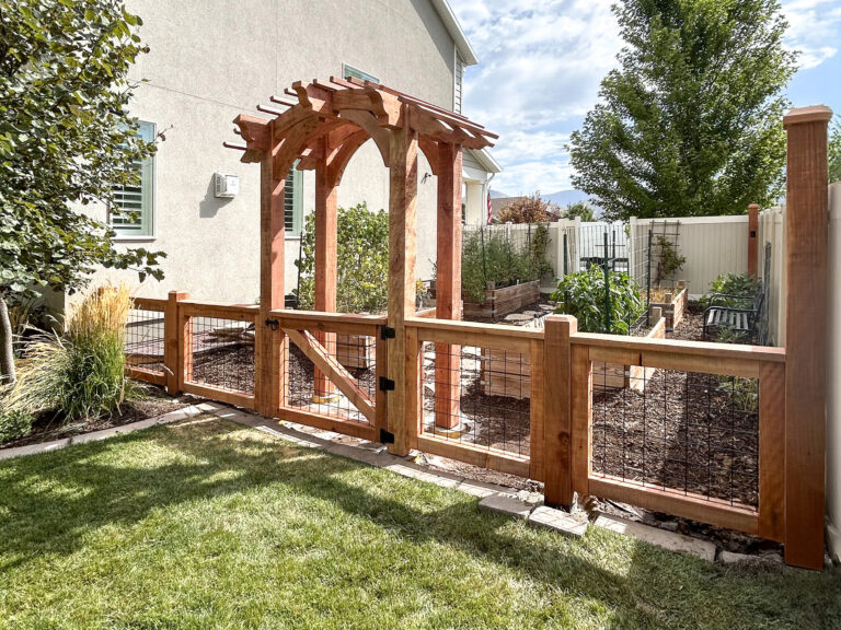 Completed Redwood arbor with matching fence and gate leading into a backyard garden with raised beds.