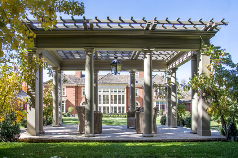 Tuscany-style timber pergola with round columns and decorative beam work, framing wicker seating in a landscaped estate garden.