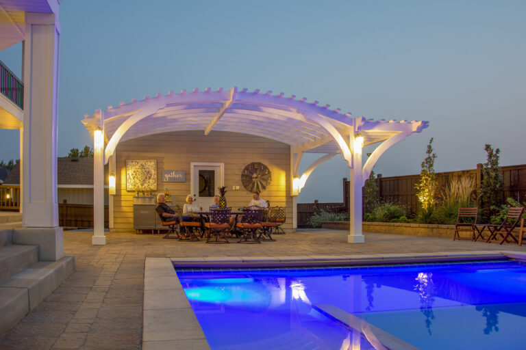 Custom arched attached timber frame pergola in white stain with cantilevered roof over poolside dining area, evening lighting, and accessible design.
