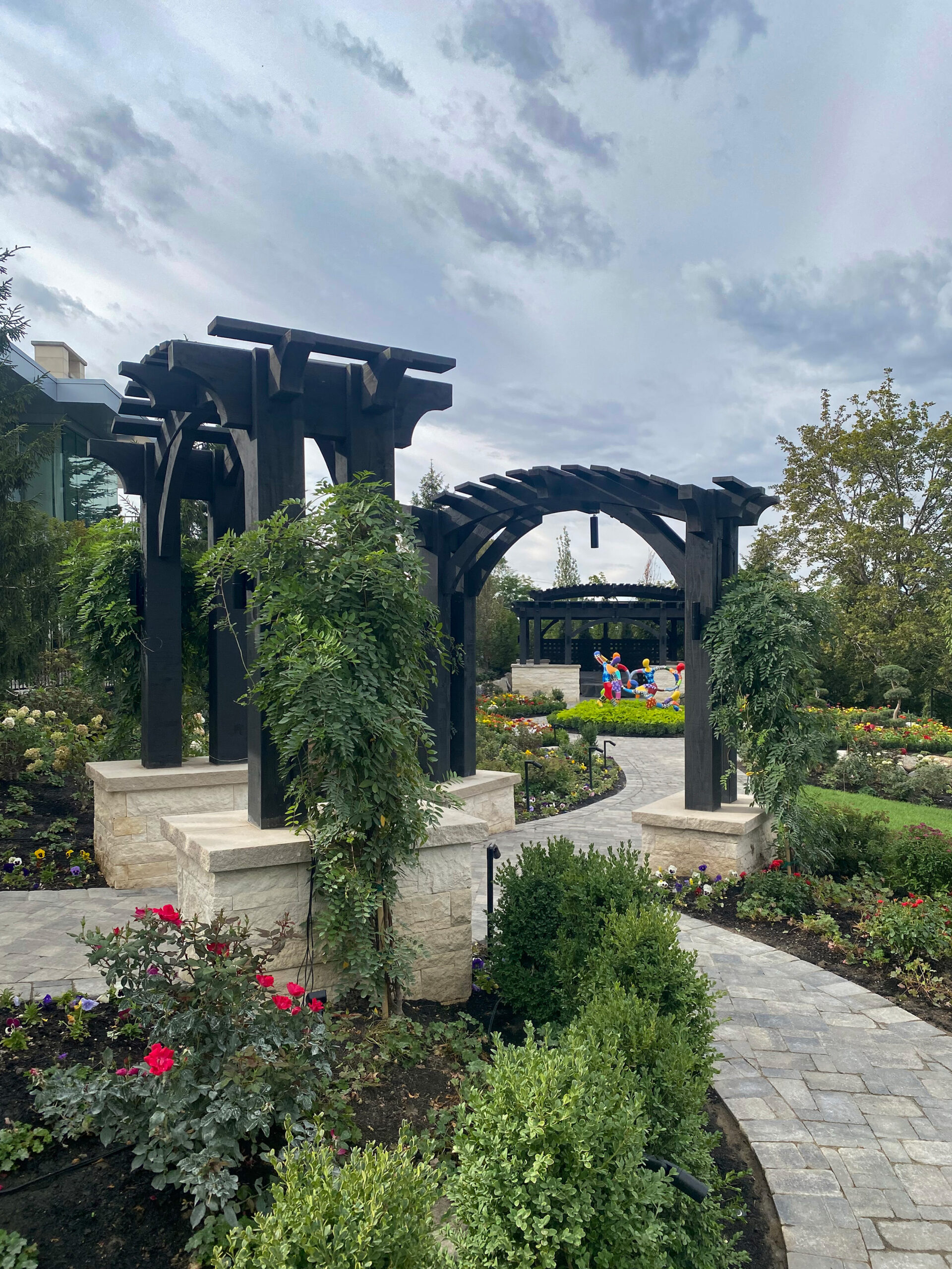 Ebony-stained timber frame arbor with stone bases and climbing vines leading to a pergola in a landscaped garden.