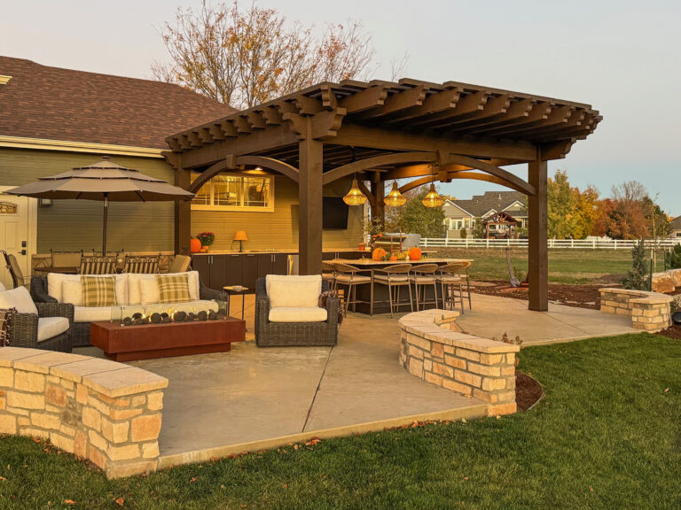 Timber frame pergola with outdoor kitchen, dining bar, and lounge seating under warm evening light in a backyard setting.