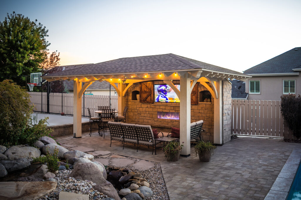 Hipped roof timber pavilion with stone fireplace, outdoor TV, and evening lighting beside a backyard pool.