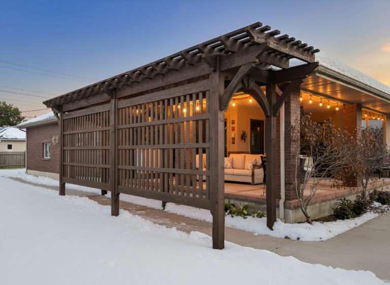 Dark stained timber pergola with lattice privacy panel attached to brick home in winter with snow on ground and warm string lights glowing inside covered patio