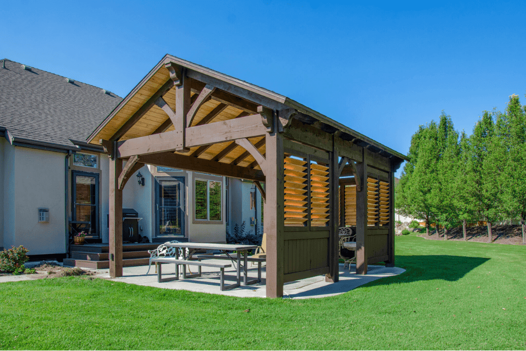 A large, dark-stained heavy timber farmhouse pavilion with a gable roof and integrated adjustable wood privacy shutters, situated on a concrete patio in a spacious green backyard.