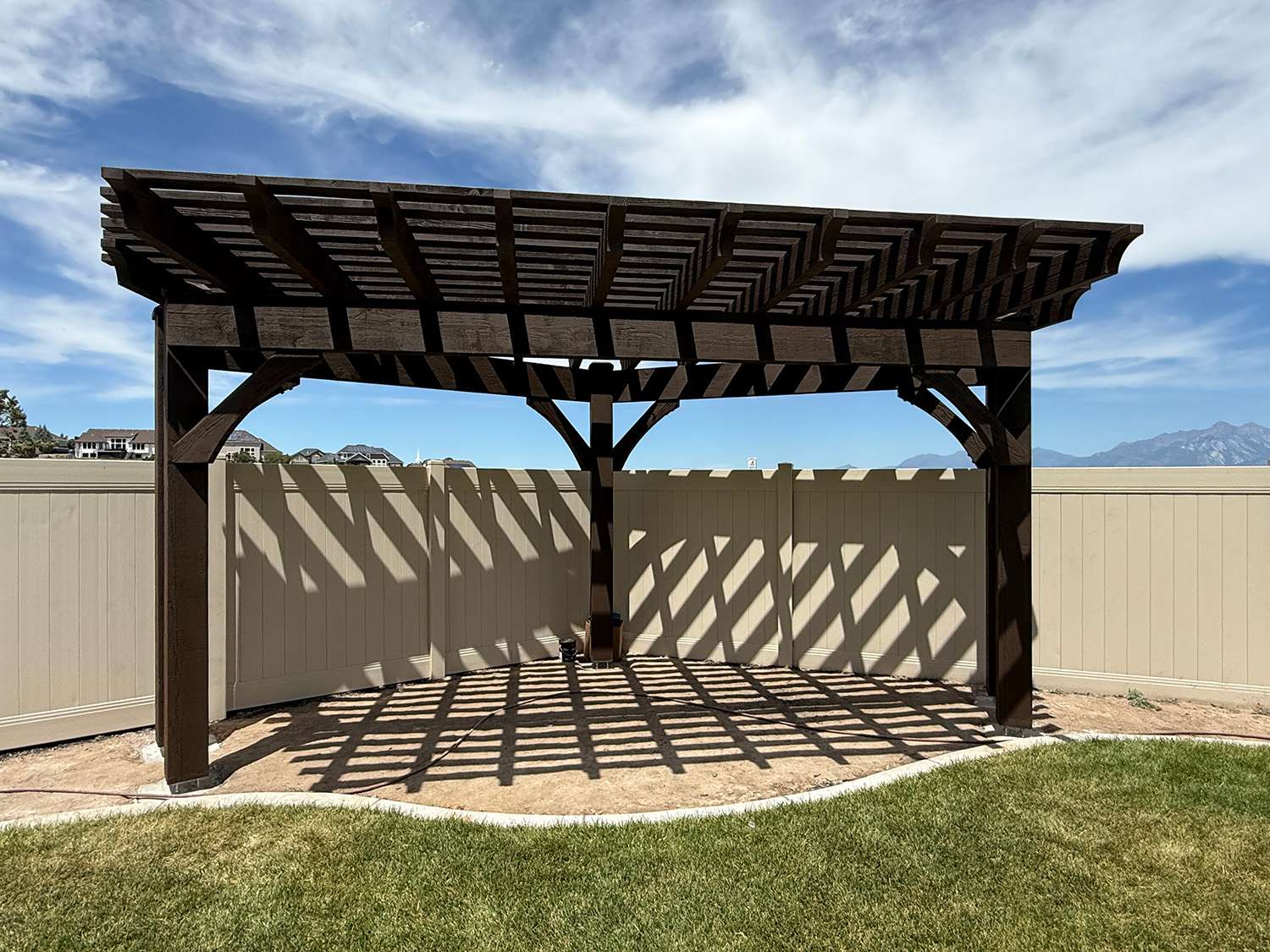 Custom five-post timber pergola with closely spaced shade planks, decorative knee braces, and dramatic shade patterns cast across a backyard patio.