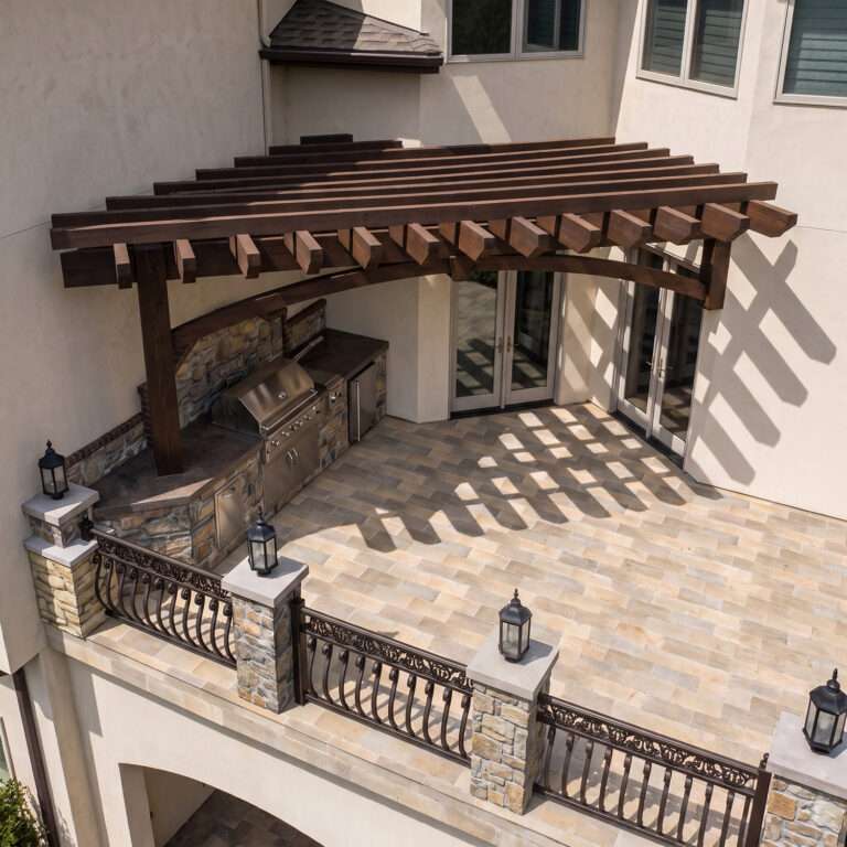 Second-story custom timber pergola covering an outdoor kitchen on an elevated patio with rough-sawn Douglas fir beams, arched braces, and luxury stone finishes.