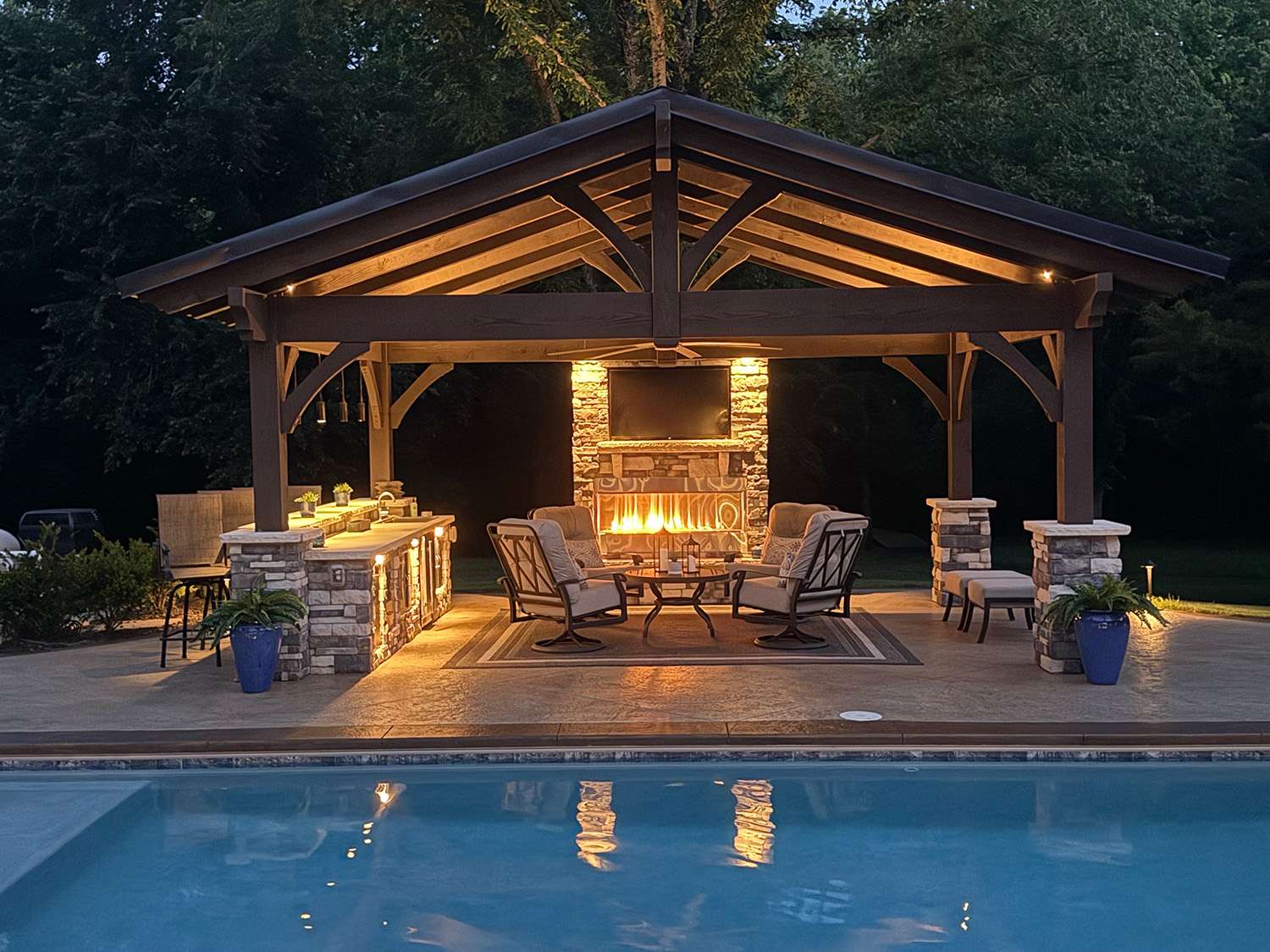 Heavy timber pavilion with Rich Cordoba stain featuring TimberVolt concealed power, outdoor kitchen, stone fireplace, TV, and poolside seating illuminated at dusk.