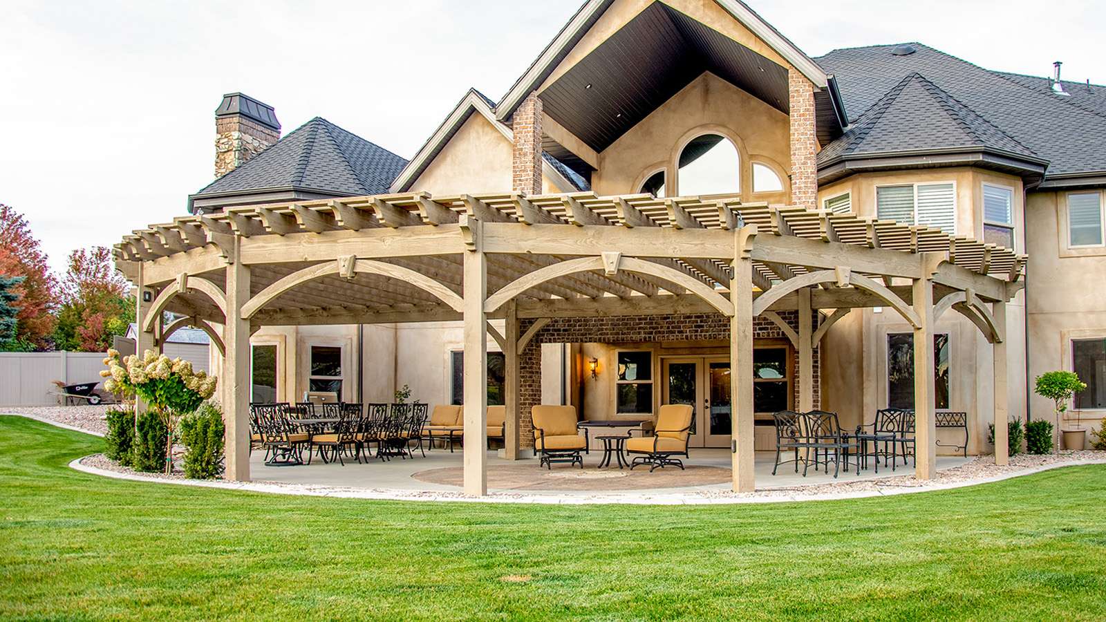 Large light-stained timber frame pergola wrapping the back of a stucco home, shading a patio with dining and lounge seating